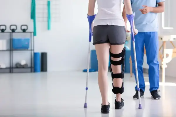 Pacjentka z ortezą na nodze uczy się chodzić o kulach pod okiem fizjoterapeuty w gabinecie rehabilitacyjnym.